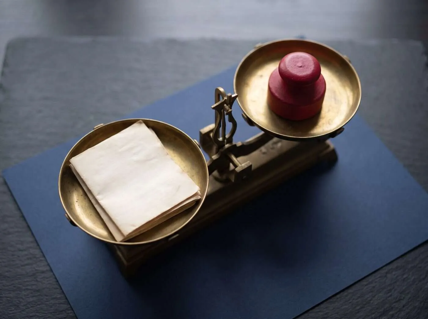 Balance en laiton sur du papier bleu marine, une pile de documents dans un plateau et un poids cramoisi dans l’autre