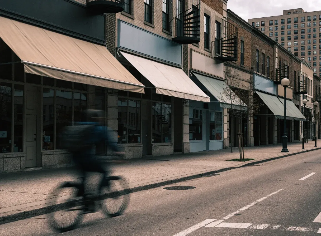 A row of independent ground-floor shops on a Montreal street, with fabric awnings, blank sign panels, spiral staircases above and a cyclist passing in motion blur