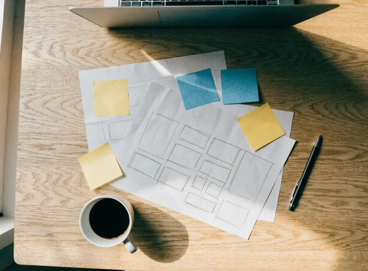 Overhead view of a website planning session: pencil wireframe sketches on paper, yellow and blue sticky notes, a pen, a cup of black coffee and the edge of an open laptop