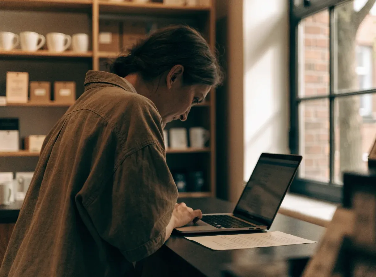 A small business owner leaning over a shop counter, reading a laptop screen with a printed document beside it