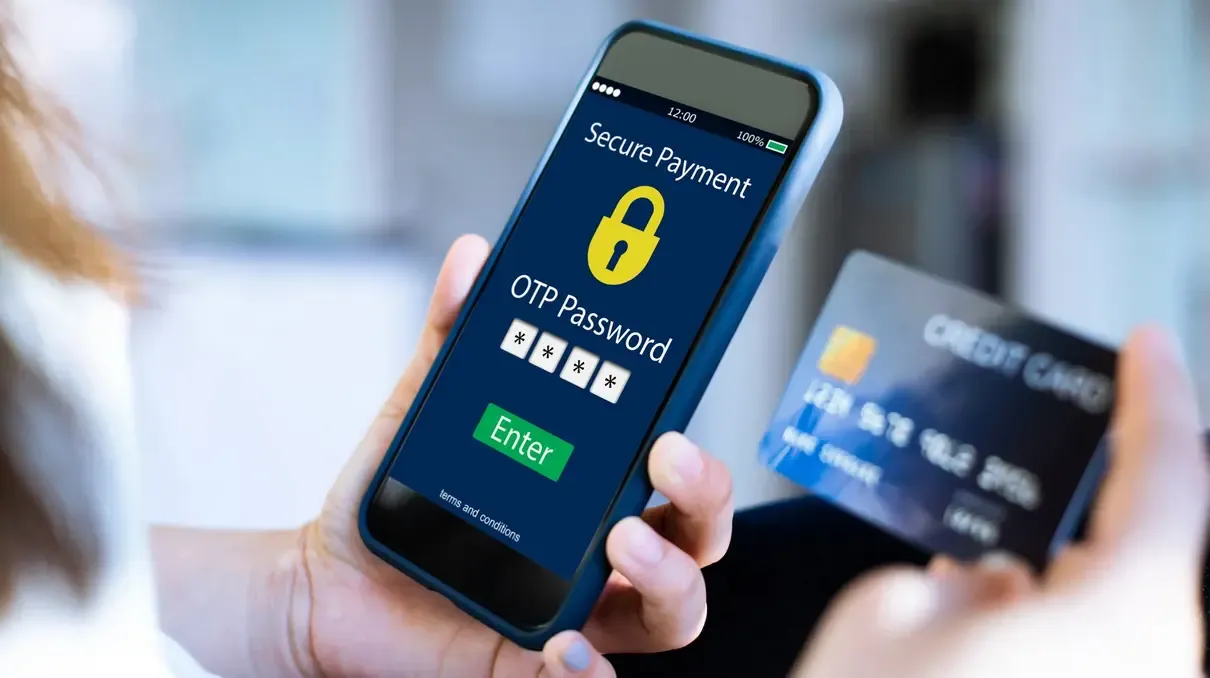 A hand holding a phone showing a secure payment screen, with a bank card held up behind it
