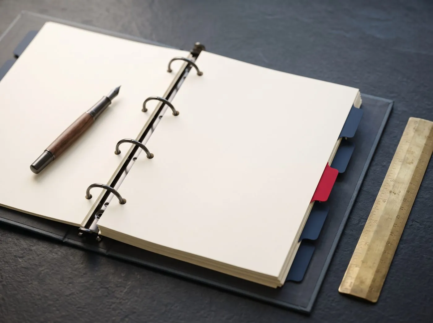 Open ring binder with navy tabbed dividers and one crimson tab, fountain pen resting on the page, brass ruler beside on a slate desk