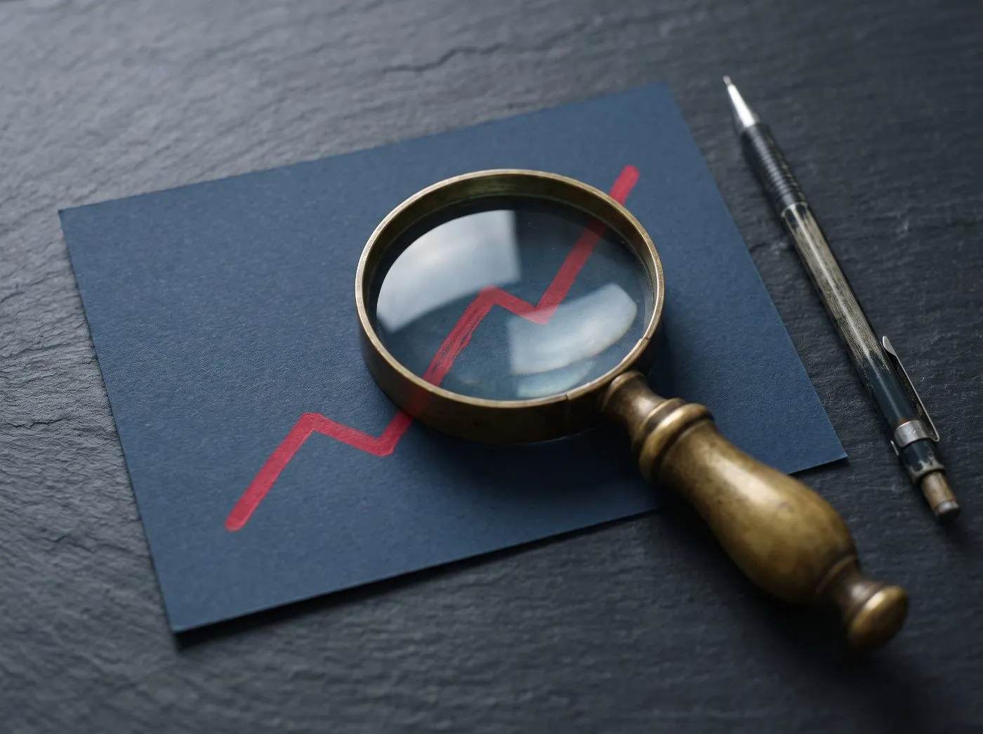 Brass magnifying glass over a crimson line chart rising across navy paper on a slate desk, mechanical pencil beside it