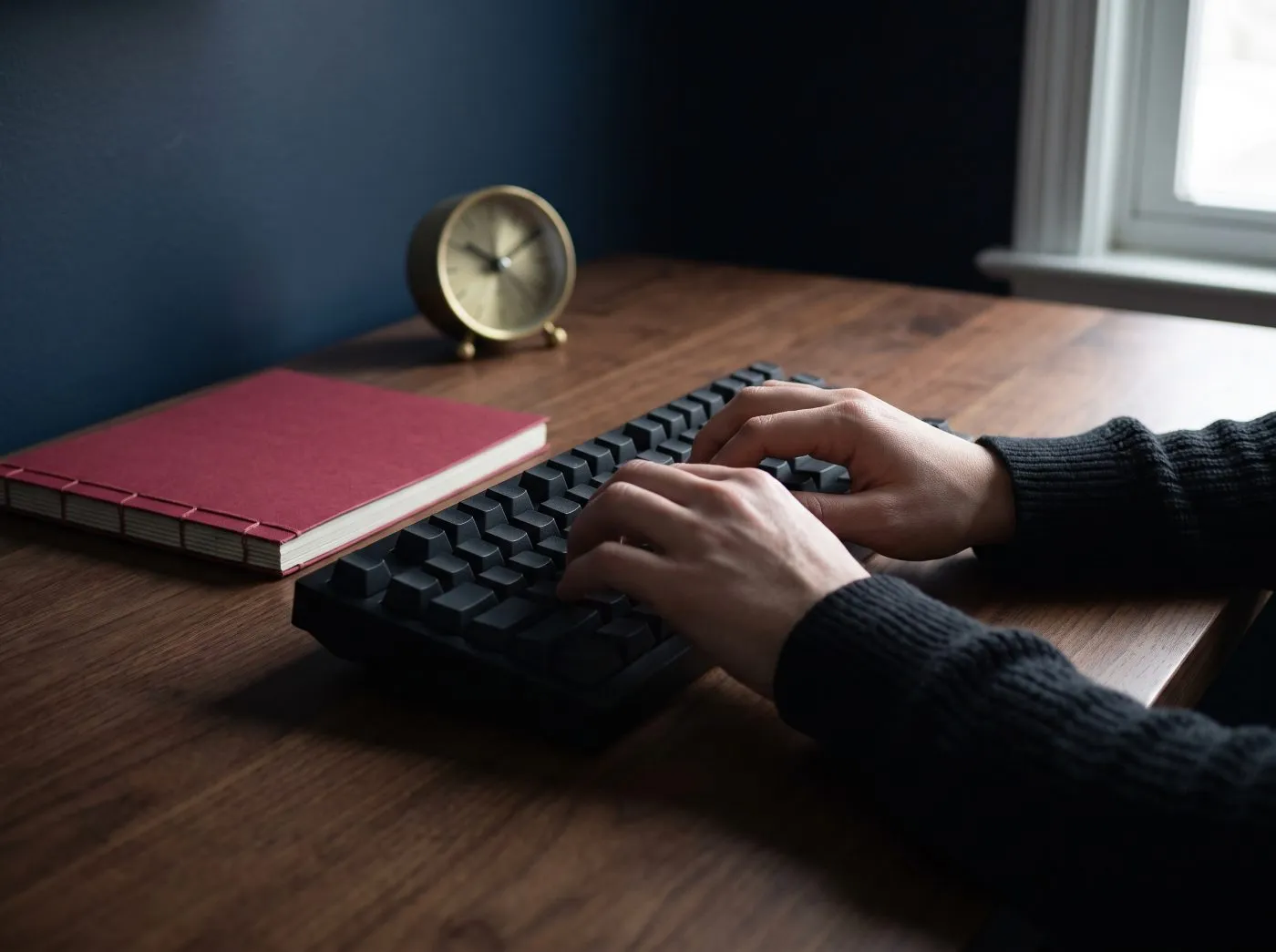 Mains d’un développeur sur un clavier mécanique noir à un bureau en noyer, à côté d’un carnet cramoisi et d’une horloge de bureau en laiton