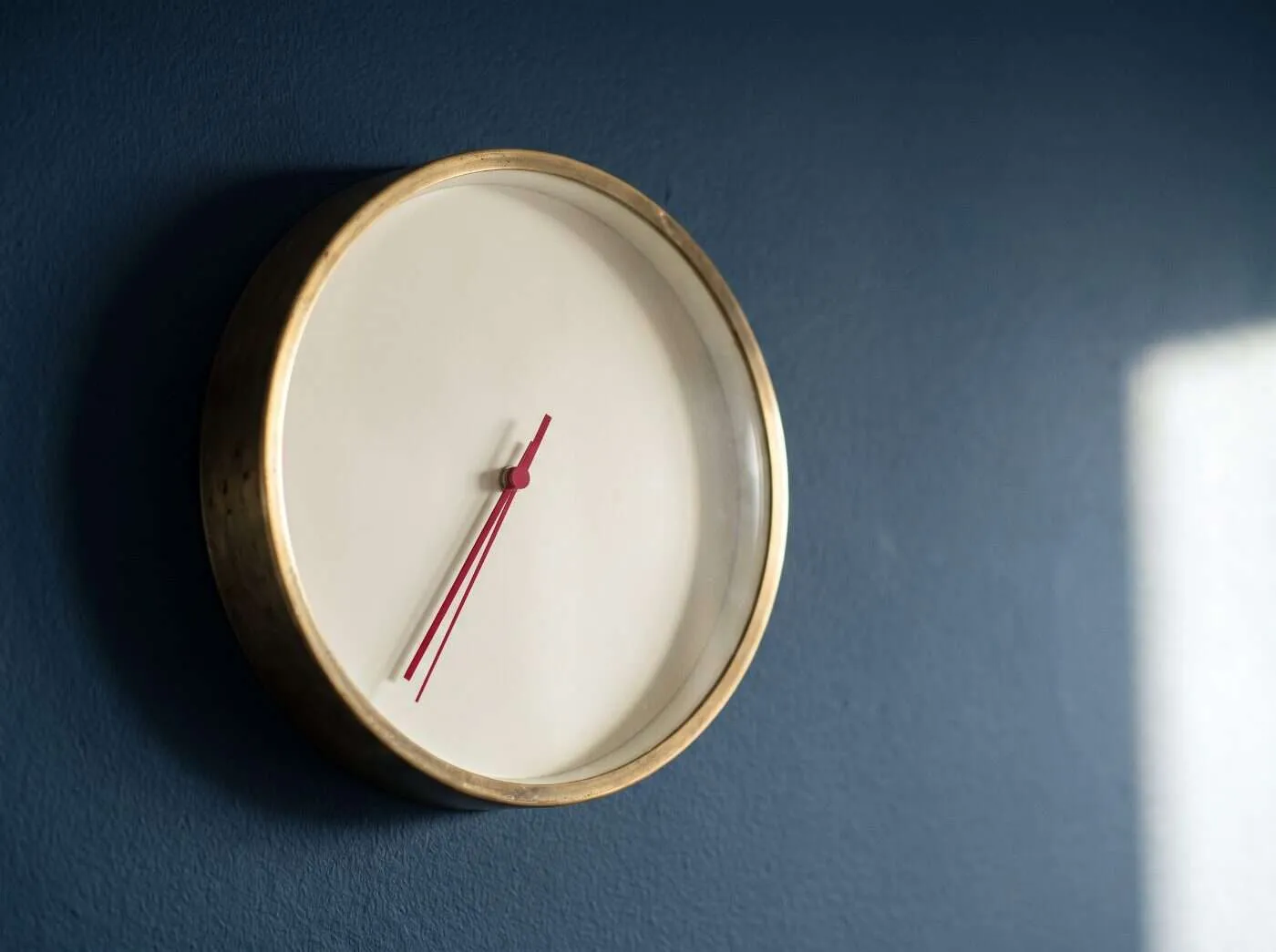 Brass-rimmed wall clock with a plain face and crimson hands on a navy wall in morning light